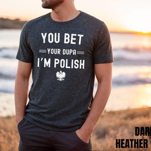 May include: A dark heather grey short-sleeved t-shirt featuring white text that reads YOU BET YOUR DUPA I'M POLISH, accompanied by a small white graphic of the Polish eagle emblem centered below the text.