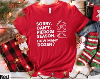 May include: A red t-shirt with white text that reads "SORRY. CAN'T. PIEROGI SEASON. HOW MANY DOZEN?" and three pierogi illustrations. The shirt is part of a holiday theme, with a gingerbread man, Christmas stocking, and festive lights.