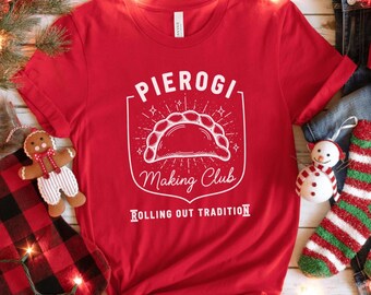 May include: A red t-shirt with a white graphic that reads "Pierogi Making Club" above an illustration of a pierogi. The text "Rolling Out Tradition" is below the graphic. The shirt is displayed with holiday decorations.