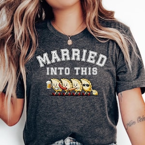 May include: Dark gray t-shirt with the text "MARRIED INTO THIS" in white block letters. Below the text is a graphic of four cartoon pierogies, each with a different accessory, walking in a line.