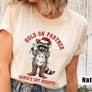 May include: A cream-colored t-shirt featuring a cartoon raccoon wearing a cowboy hat and Santa hat, holding a candy cane and wrapped in Christmas lights. The text "HOLD ON PARTNER" and "SANTA'S GOT SCRIPTS" are printed in red.