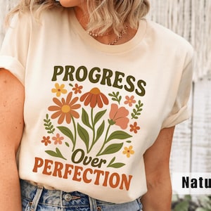 Puede incluir: Una camiseta de color natural con las palabras "PROGRESS Over PERFECTION" en marrón y naranja. La camiseta presenta un diseño floral con flores naranjas, melocotón y amarillas con hojas verdes.