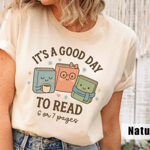 May include: A cream-colored t-shirt with the text "It's a good day to read 6 or 7 pages." The graphic features three cartoon books with smiling faces, a coffee cup, and a scarf. The shirt is labeled "Natural".