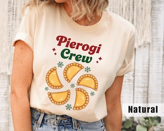 May include: A cream-colored t-shirt with the words "Pierogi Crew" in red and green, with a design of pierogies and snowflakes. The shirt is a casual style, perfect for food lovers.