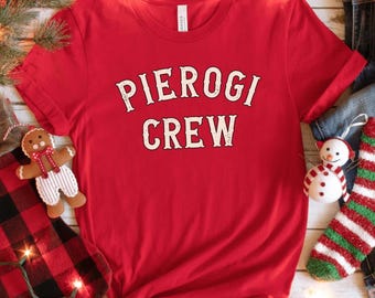 May include: A red t-shirt with the words "PIEROGI CREW" in a white, vintage-style font. The shirt is part of a holiday-themed display, with a gingerbread man, a snowman ornament, and a red and green striped stocking.