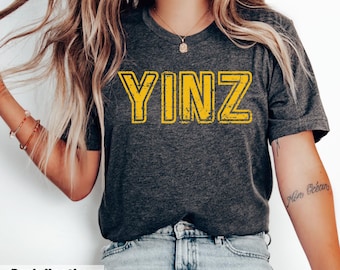 May include: A dark heather grey short-sleeve t-shirt featuring the word YINZ printed in a distressed yellow font across the chest. The shirt has a crew neckline and is paired with light blue denim jeans, a simple necklace, and bracelets.