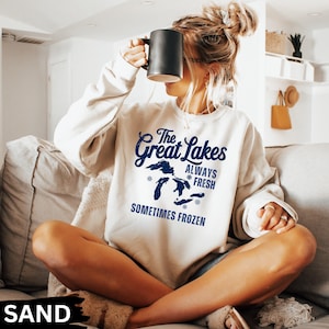 May include: A sand-coloured sweatshirt featuring the navy blue text "The Great Lakes ALWAYS FRESH SOMETIMES FROZEN" and a map of the Great Lakes. A black mug is held up to the face.