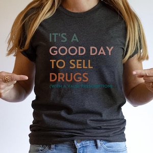 May include: A dark charcoal gray short-sleeve t-shirt featuring the text IT'S A GOOD DAY TO SELL DRUGS (WITH A VALID PRESCRIPTION) printed in stacked lines of muted green, light pink, yellow, and orange lettering across the chest area.