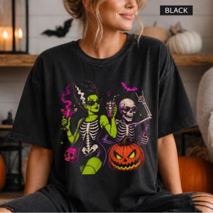 May include: A black t-shirt featuring a graphic print of a green-skinned character with a tall hairstyle and a skeleton, both wearing sunglasses. The design includes a jack-o'-lantern, a pink martini glass, a skull-shaped bag, and purple bats.