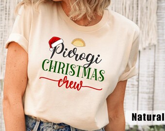 May include: A natural-colored t-shirt with the words "Pierogi Christmas Crew" in black, green, and red script. The design includes a Santa hat and a pierogi illustration. The shirt is a casual, festive apparel item.