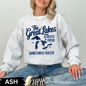 May include: A light grey sweatshirt featuring the navy blue text "The Great Lakes ALWAYS FRESH SOMETIMES FROZEN". The design includes a map of the Great Lakes and snowflake graphics. The model is wearing blue jeans and black and white trainers.