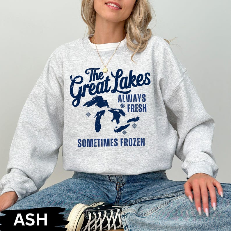 May include: A light grey sweatshirt featuring the navy blue text "The Great Lakes ALWAYS FRESH SOMETIMES FROZEN". The design includes a map of the Great Lakes and snowflake graphics. The model is wearing blue jeans and black and white trainers.
