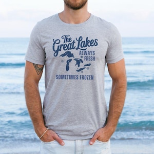 May include: A grey t-shirt featuring the text "The Great Lakes Always Fresh Sometimes Frozen" in navy blue, with a map of the Great Lakes. The shirt is worn by a person. The background is the sea.