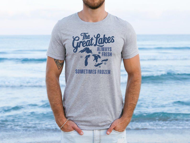 May include: A grey t-shirt featuring the text "The Great Lakes Always Fresh Sometimes Frozen" in navy blue, with a map of the Great Lakes. The shirt is worn by a person. The background is the sea.