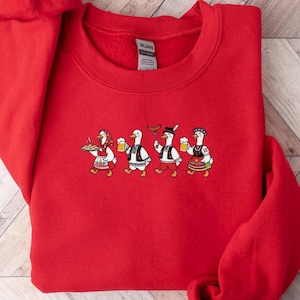 May include: Red sweatshirt featuring a cartoon design of four geese in traditional outfits. One goose carries a tray of food, others hold beer steins and a sausage. The geese are in a line, with the design centered on the chest.