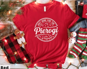 May include: A red t-shirt with the text "JUST HERE FOR THE Pierogi" in a white circular design. The shirt is displayed with holiday decorations, including a gingerbread man, a Christmas stocking, and a snowman.