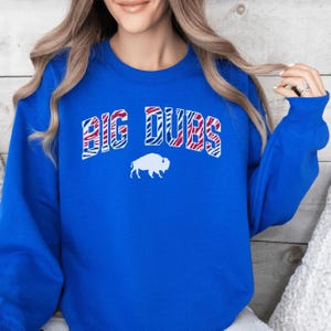 May include: Royal blue sweatshirt with the text "BIG DUBS" in red, white, and blue patterned lettering. A white buffalo silhouette is centered below the text. The sweatshirt is a crew neck style.