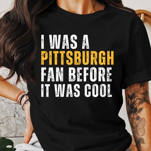 May include: Black t-shirt with the text "I WAS A PITTSBURGH FAN BEFORE IT WAS COOL" in white and yellow lettering. The shirt is a classic crew neck style.