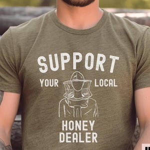 May include: Olive green t-shirt with the text "SUPPORT YOUR LOCAL HONEY DEALER" in white block letters. A line drawing of a beekeeper in protective gear is below the text. The shirt is made of a soft material.
