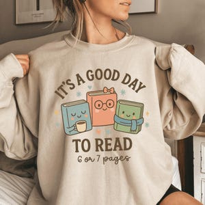 May include: Beige sweatshirt with the text "It's a good day to read 6 or 7 pages." The graphic features three cartoon books: a blue book holding a coffee cup, a pink book with glasses, and a green book with a scarf.