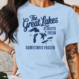 May include: Pale blue t-shirt with navy blue text: "The Great Lakes ALWAYS FRESH SOMETIMES FROZEN". The design includes a map of the Great Lakes and snowflakes. The shirt is made of a soft material.