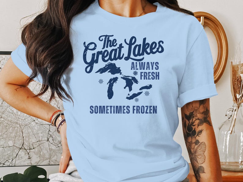 May include: Pale blue t-shirt with navy blue text: "The Great Lakes ALWAYS FRESH SOMETIMES FROZEN". The design includes a map of the Great Lakes and snowflakes. The shirt is made of a soft material.