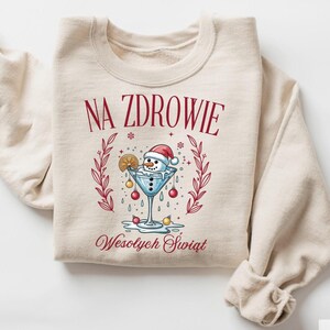 May include: Beige sweatshirt with a festive design. The graphic features a snowman in a martini glass, wearing a Santa hat, surrounded by ornaments. The text "NA ZDROWIE" and "Wesołych Świąt" are printed in red.