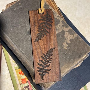 May include: A wooden bookmark with a fern design, featuring a brown leather tassel. The bookmark is made of dark wood and has a natural finish.