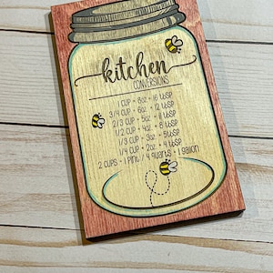 May include: A wooden kitchen conversion chart in the shape of a mason jar with a bee design. The chart lists common kitchen conversions for cups, ounces, and tablespoons.