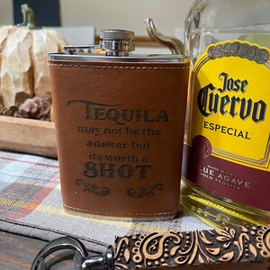 May include: A brown leather-wrapped flask with the text "TEQUILA may not be the answer but its worth a SHOT" engraved on the front. The flask is next to a bottle of Jose Cuervo Especial Tequila and a wooden bottle opener with a paisley design.