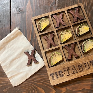 May include: Wooden tic-tac-toe game with a taco theme. The game board is made of wood and has a brown stain. The game pieces are made of wood and are shaped like tacos. The game pieces are painted with yellow, brown, and green paint. The game board has the words "Tic-Tac-Toe" written on it.