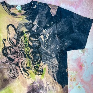 Op de afbeelding: Een zwart T-shirt met een tie-dye patroon en een groene en zwarte octopus grafische print.