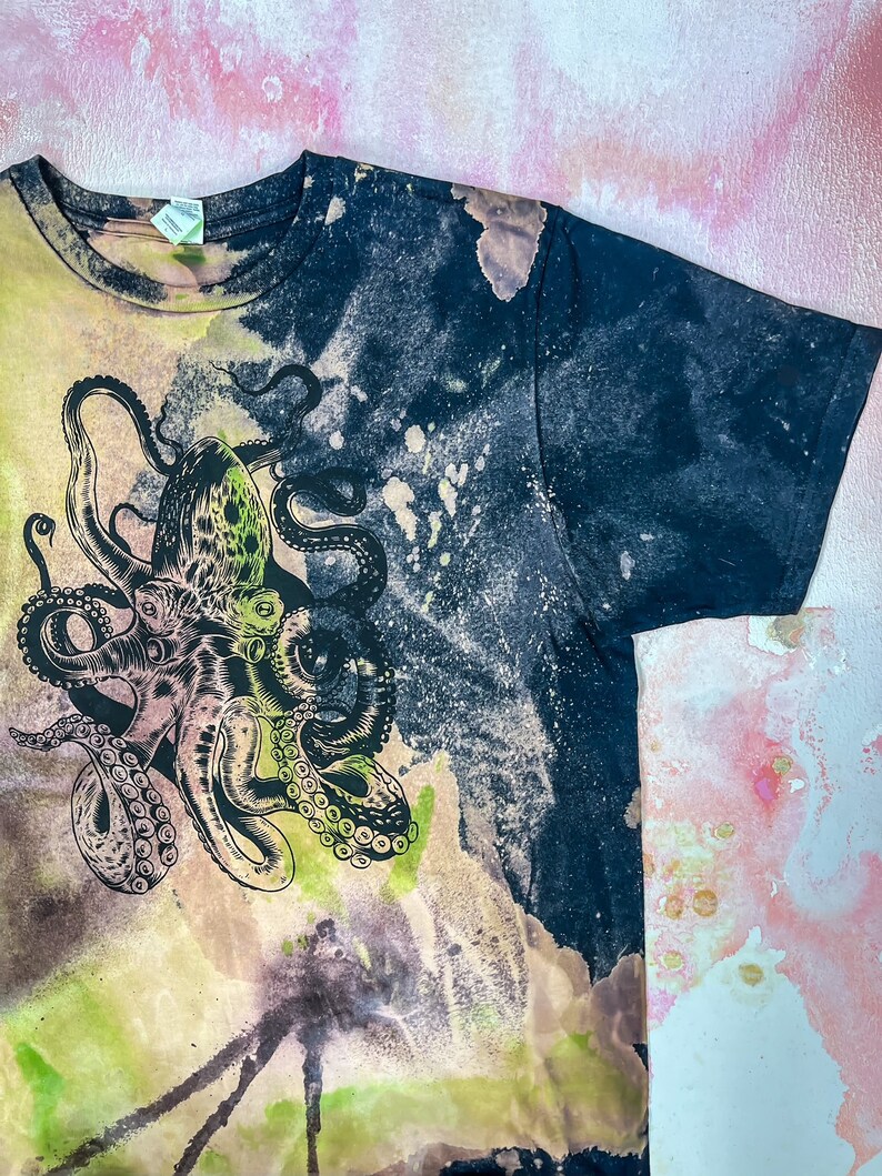 Op de afbeelding: Een zwart T-shirt met een tie-dye patroon en een groene en zwarte octopus grafische print.