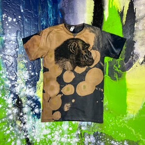 May include: A black and brown t-shirt with a bleach-splattered design. The shirt features a black and white image of a chimpanzee's head in profile.