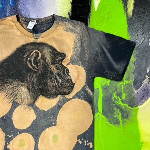 May include: A black t-shirt with a bleach-splattered design featuring a black and white illustration of a chimpanzee's face. The shirt has a brown, faded look.