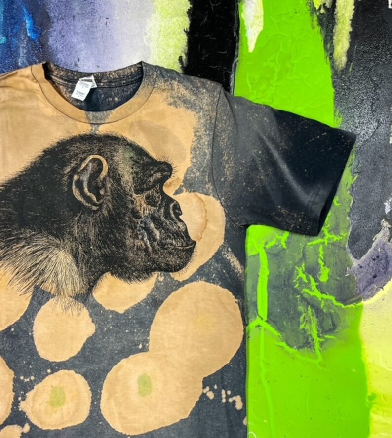 May include: A black t-shirt with a bleach-splattered design featuring a black and white illustration of a chimpanzee's face. The shirt has a brown, faded look.