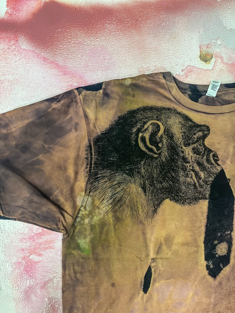 May include: A brown t-shirt with a black and white image of a chimpanzee's face on the front.