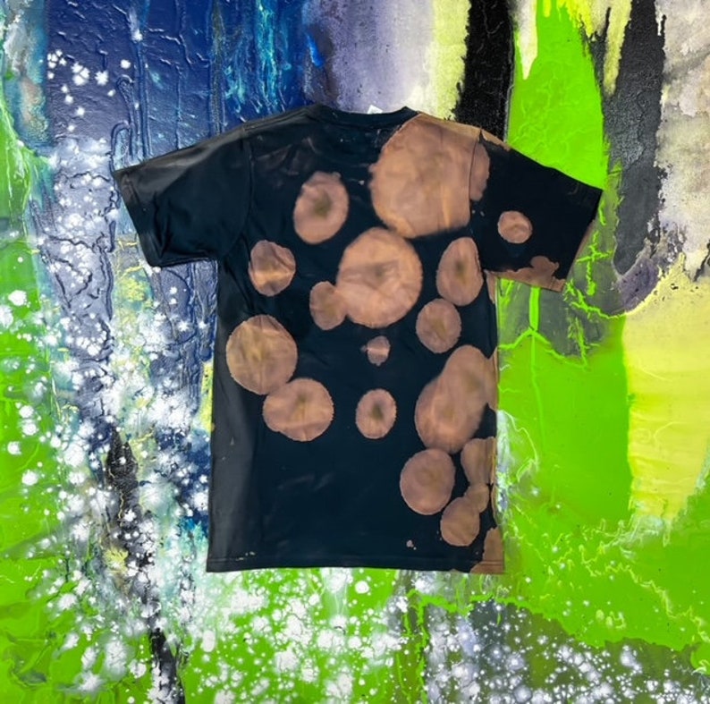 May include: A black t-shirt with a bleach-splattered design. The design features a pattern of irregular circles in shades of brown.