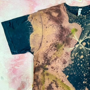 May include: A black t-shirt with a tie-dye pattern in shades of brown, yellow, and green. The pattern resembles a galaxy with stars and a swirling design.