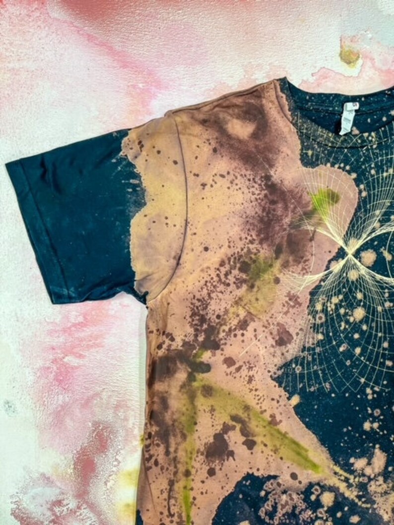 May include: A black t-shirt with a tie-dye pattern in shades of brown, yellow, and green. The pattern resembles a galaxy with stars and a swirling design.