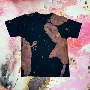 May include: Black t-shirt with brown bleach splatters and a faded design. The shirt has a crew neck and short sleeves.