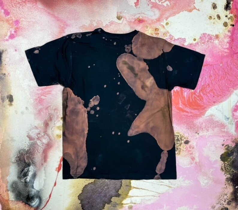 May include: Black t-shirt with brown bleach splatters and a faded design. The shirt has a crew neck and short sleeves.