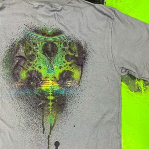 May include: Gray t-shirt with a green, blue, and black spray paint design of a swirling, abstract pattern.