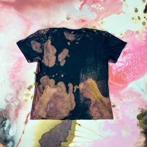 K&ouml;nnte beinhalten: Ein schwarzes T-Shirt mit einem Bleichmittel-Spritzer-Design. Das Shirt hat einen verblassten, abgenutzten Look mit braunen und violetten Flecken.