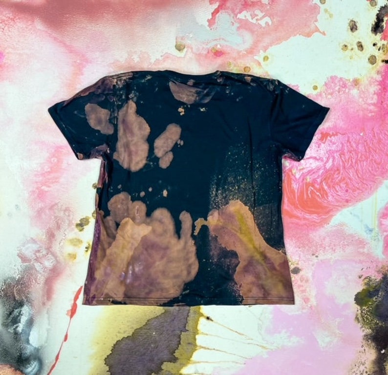 K&ouml;nnte beinhalten: Ein schwarzes T-Shirt mit einem Bleichmittel-Spritzer-Design. Das Shirt hat einen verblassten, abgenutzten Look mit braunen und violetten Flecken.