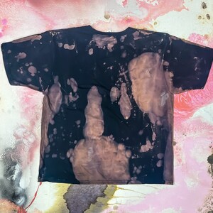 May include: A black t-shirt with a bleach-splattered design. The shirt has a distressed look with brown and white splotches.