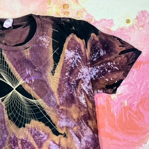 K&ouml;nnte beinhalten: Ein schwarzes T-Shirt mit einem Batik-Muster in Violett-, Braun- und Wei&szlig;t&ouml;nen. Das Shirt hat ein geometrisches Design mit einem wei&szlig;en Umriss.