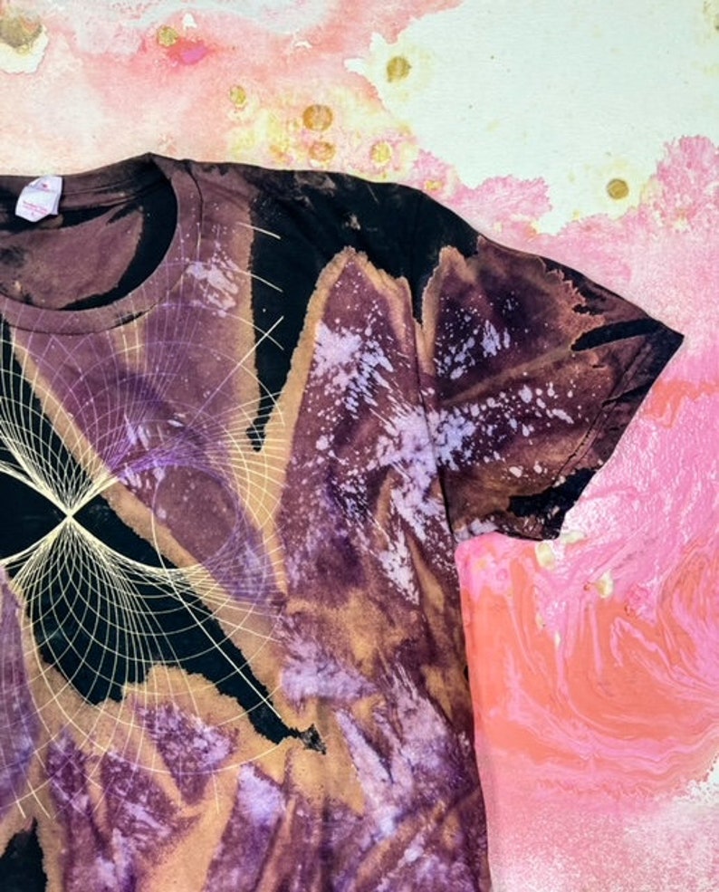 K&ouml;nnte beinhalten: Ein schwarzes T-Shirt mit einem Batik-Muster in Violett-, Braun- und Wei&szlig;t&ouml;nen. Das Shirt hat ein geometrisches Design mit einem wei&szlig;en Umriss.