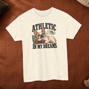 Könnte beinhalten: Ein cremefarbenes Kurzarm-T-Shirt mit einer Grafik eines Französischen Bulldoggen, der auf einem grünen Kissen mit einer roten Hantel liegt. Über dem Hund steht ATHLETIC, darunter IN MY DREAMS. Eine Sprechblase zeigt einen Sportschuh.