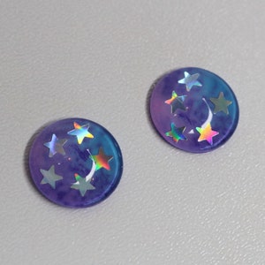 Op de afbeelding: Een paar ronde hars oorbellen met een paars en blauw ombre-effect. De oorbellen zijn versierd met iriserende holografische sterren.